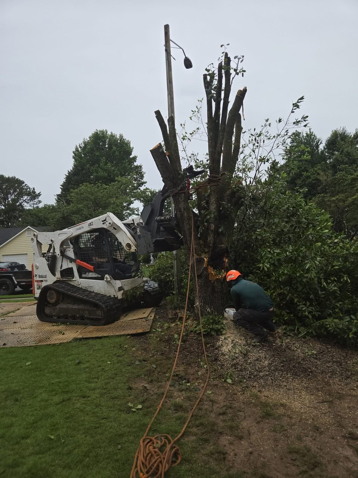 Broken trunk rigged to a skid steer while a climber cuts it at the base in Acworth GA, June 2026
