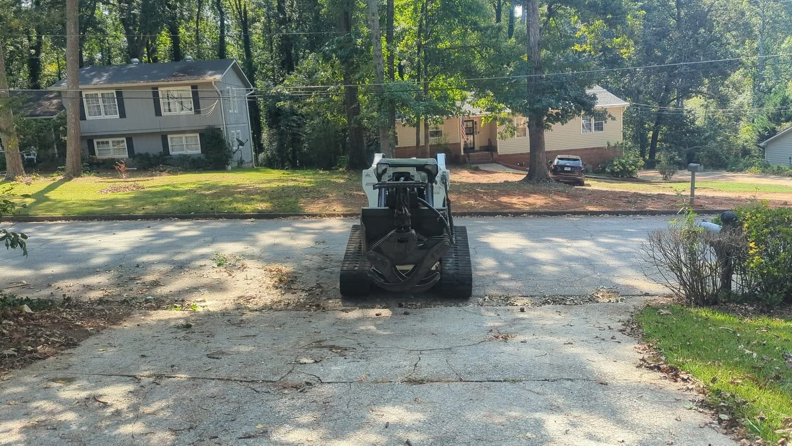 Tracked skid steer with log grapple staged on a residential driveway during storm cleanup