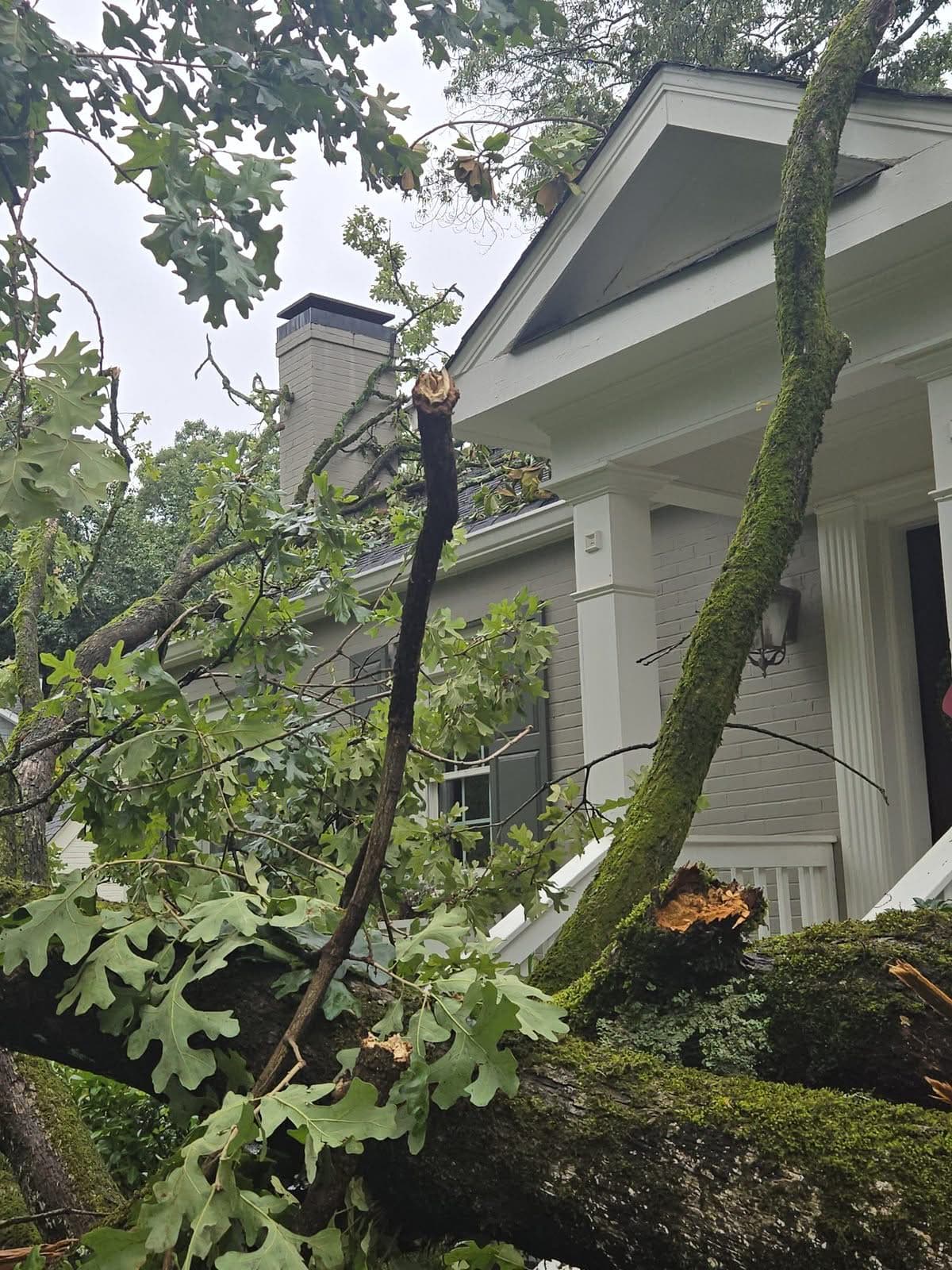 Broken oak limbs resting over a home's front porch roofline with a chimney behind