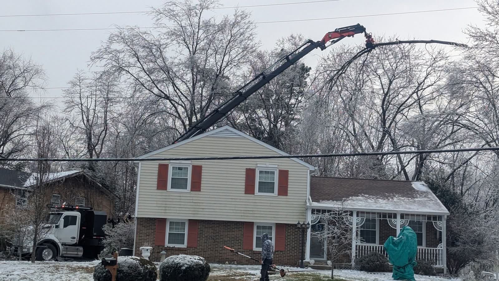 Red Palfinger knuckleboom crane parked in front of a house, boom arched over the roof to grip a limb in ice-covered trees