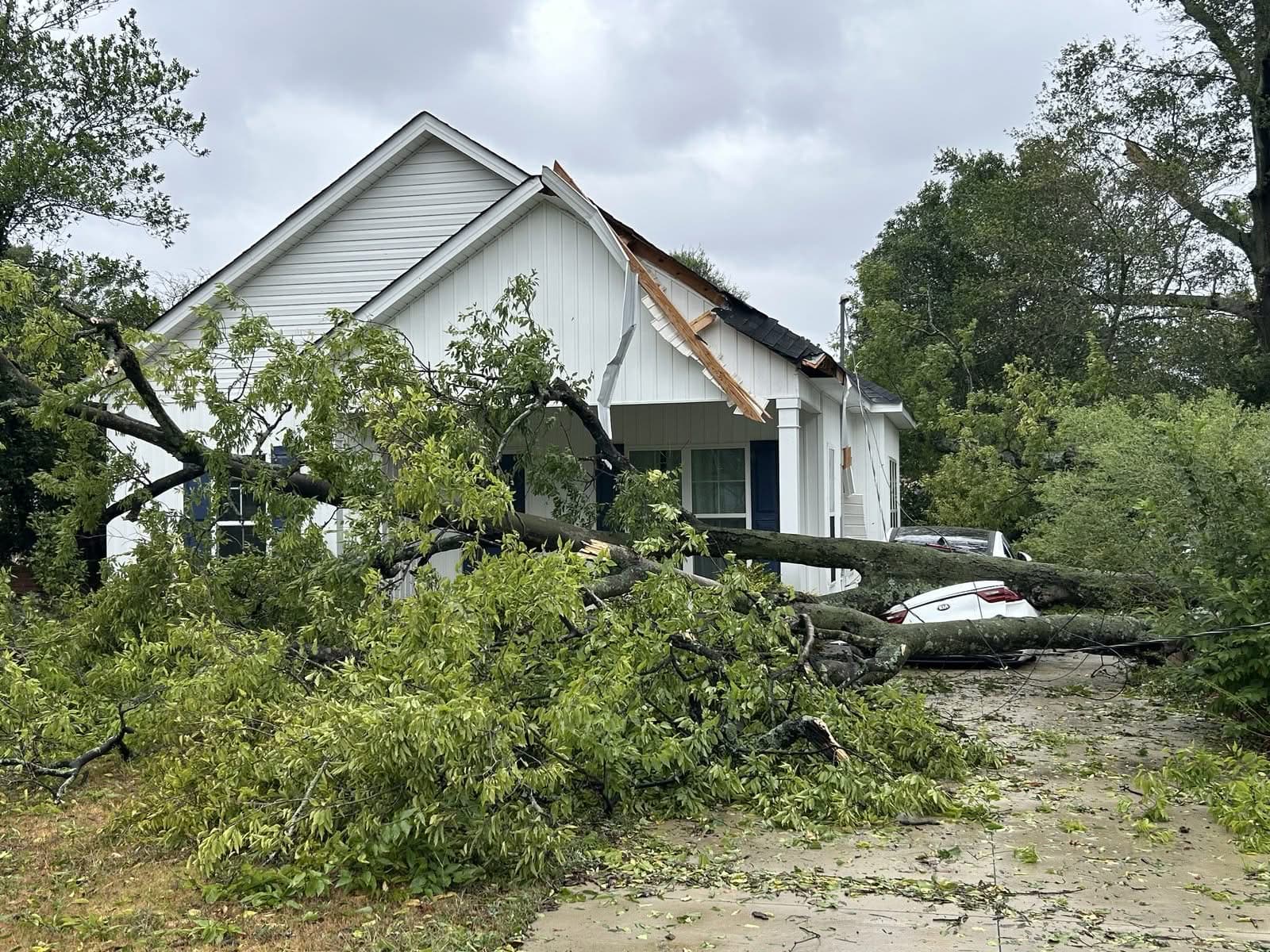 Storm-fallen tree across a front yard, its trunk crushing a car in the driveway, with the house's porch gable torn open behind it