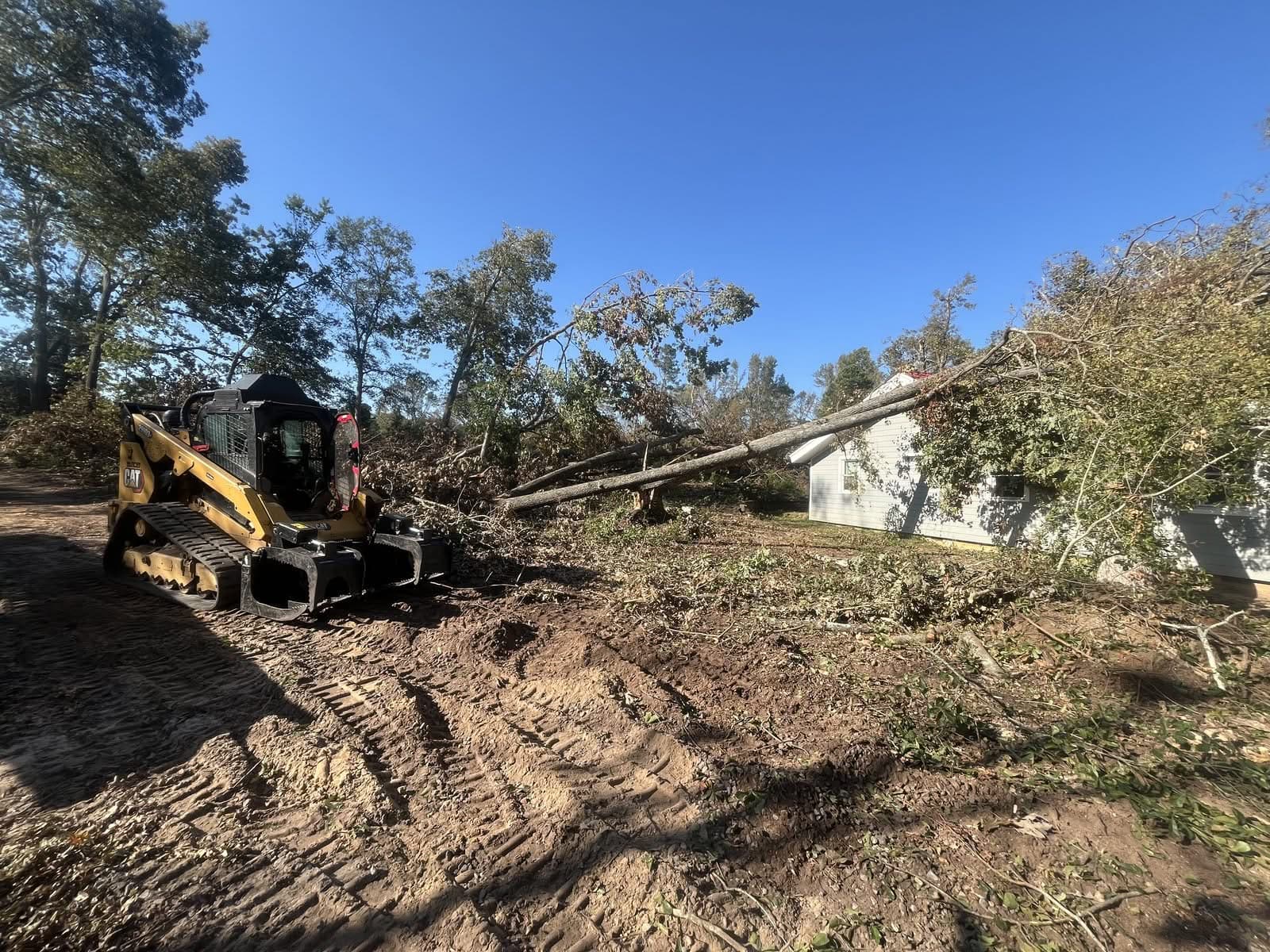 Tracked skid steer with a grapple on a storm site where a fallen tree rests against a house