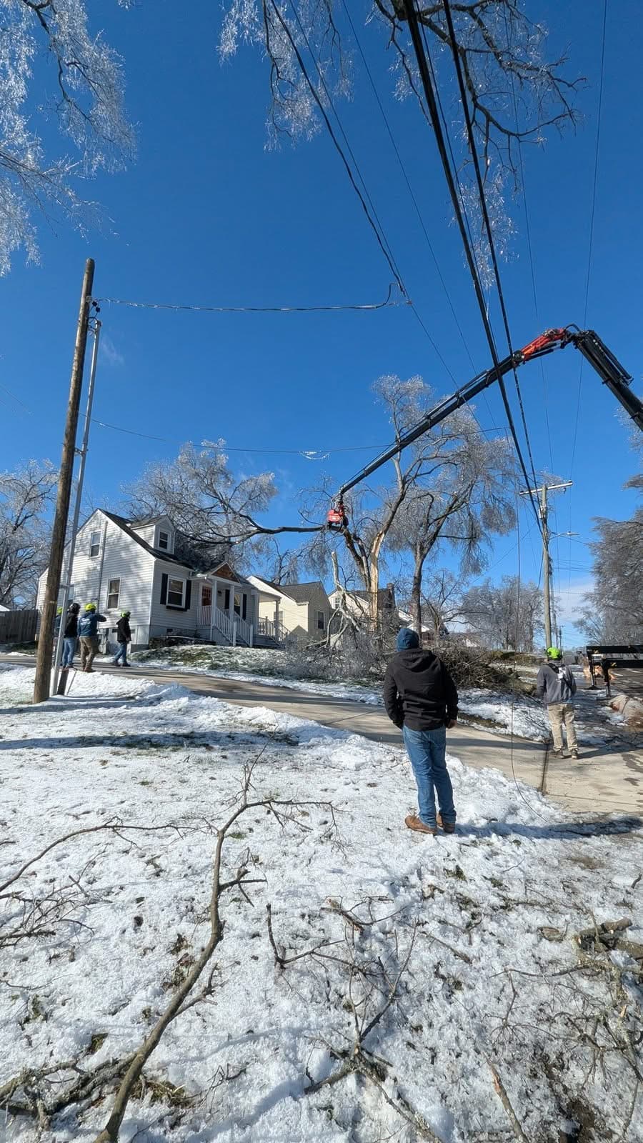 Knuckleboom crane boom extended over a snowy street toward a split, ice-coated tree, crew standing back near the power lines